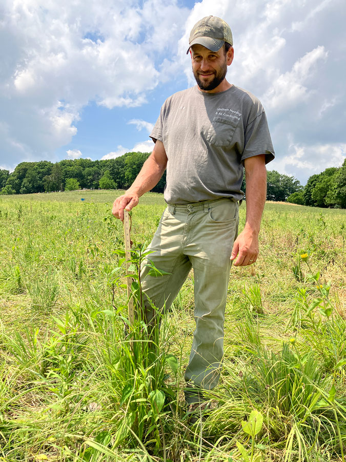 A white man with work clothes and a baseball cap, standing in a prairie pasture. He is holding a measuring stick next to a tall plant.