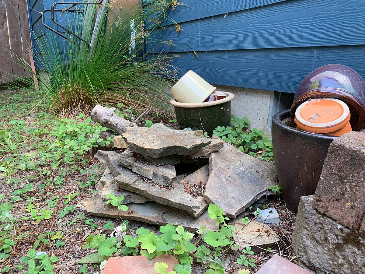 A small rock pile in a yard, alongside the side of a house. Made of flat stones stacked together, gaps have been left to allow for animals to move inside the pile. Plants grow alongside the pile.