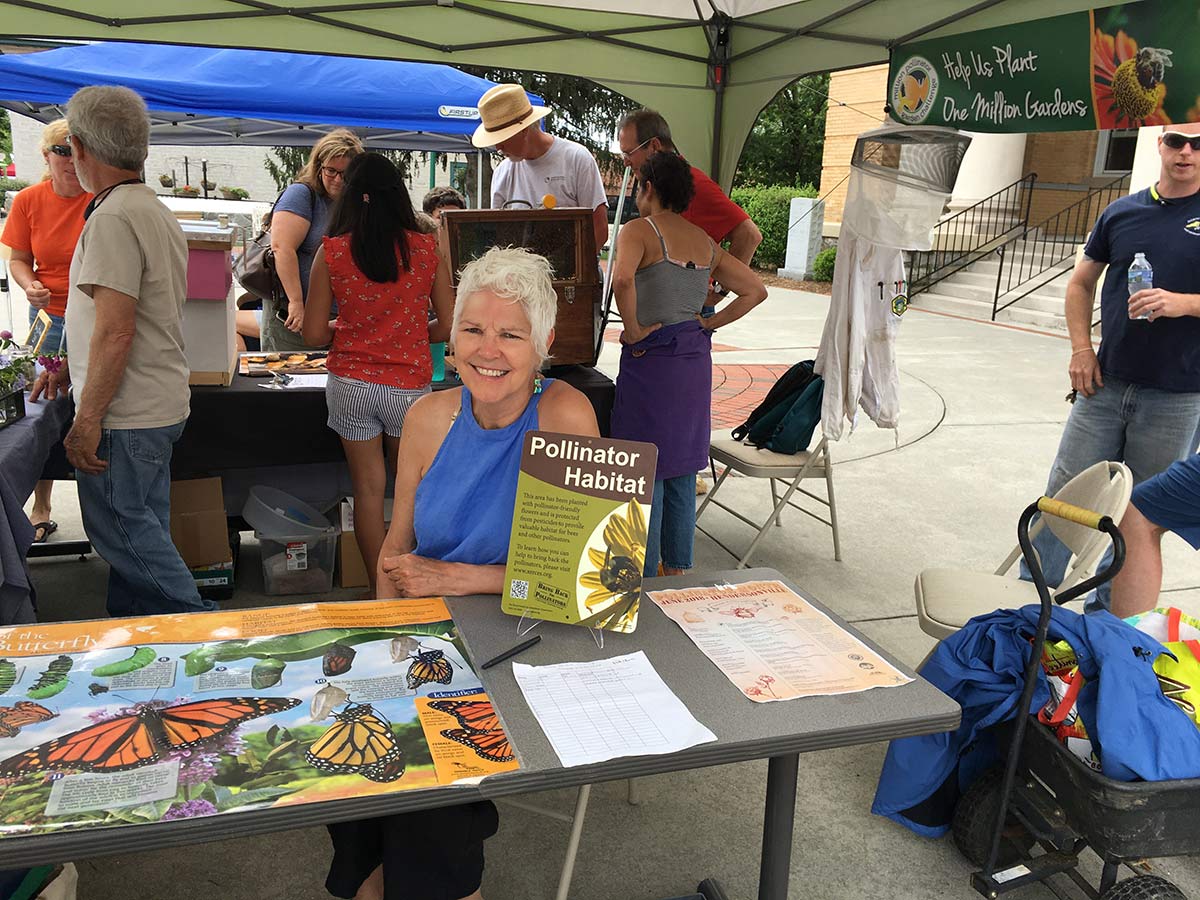  Phyllis Stiles, an older white woman with grey hair, presenting a table display at a local environmental festival. She is smiling and surrounded by information about pollinators. 