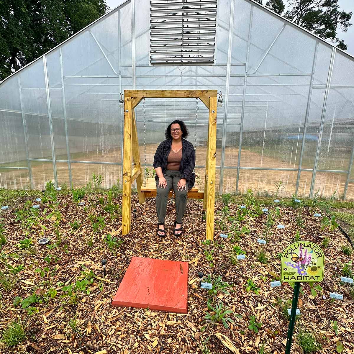 Stefanie, a black woman and mixed race with shoulder length curly hair and glasses, smiling and sitting on a bench in front of a newly installed pollinator habitat. Behind her is a structure for growing crops.. 