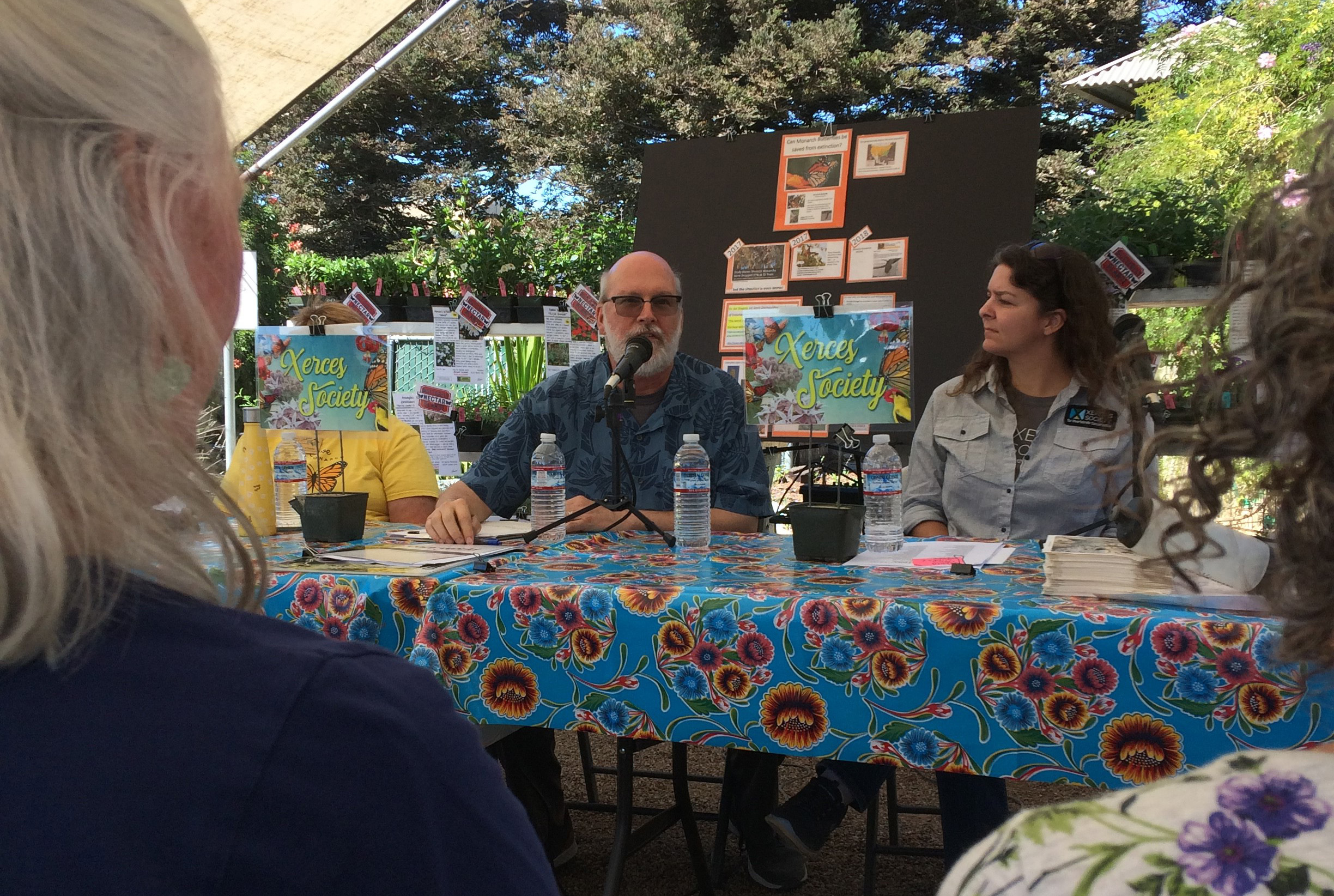 A bearded man sits behind a table speaking to a small gathering of gardeners.