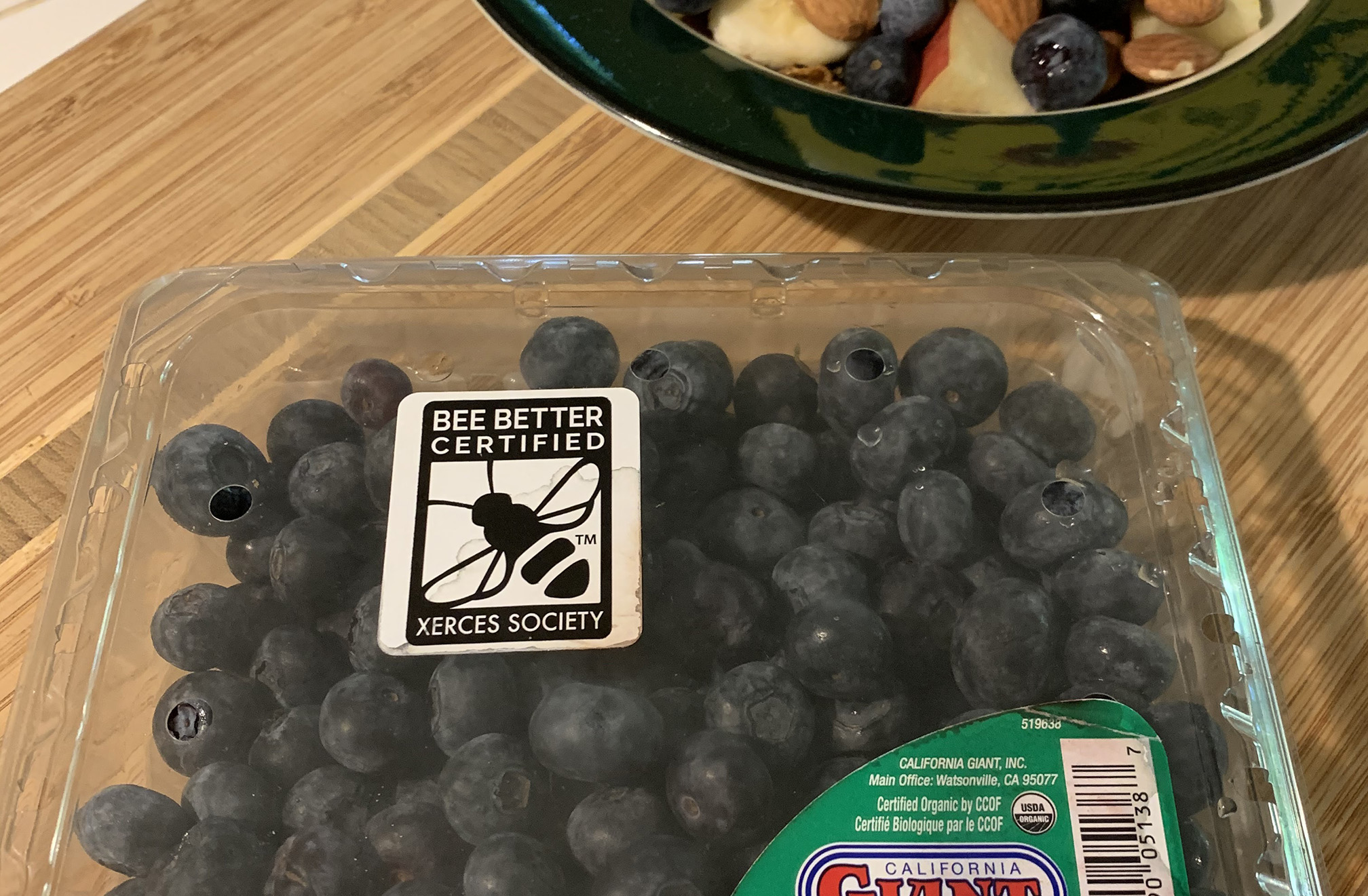 A clear plastic container of blueberries on a kitchen counter beside a breakfast bowl containing cereal and fruit. The blueberry container has the black-and-white Bee Better Certified label
