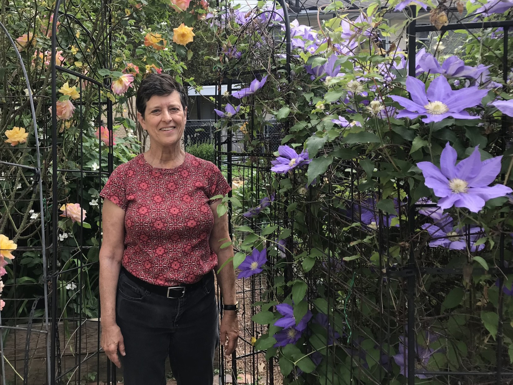 Jan Mooney stands beside purple clematis and yellow rose vines at Upper Fells Point Community Garden