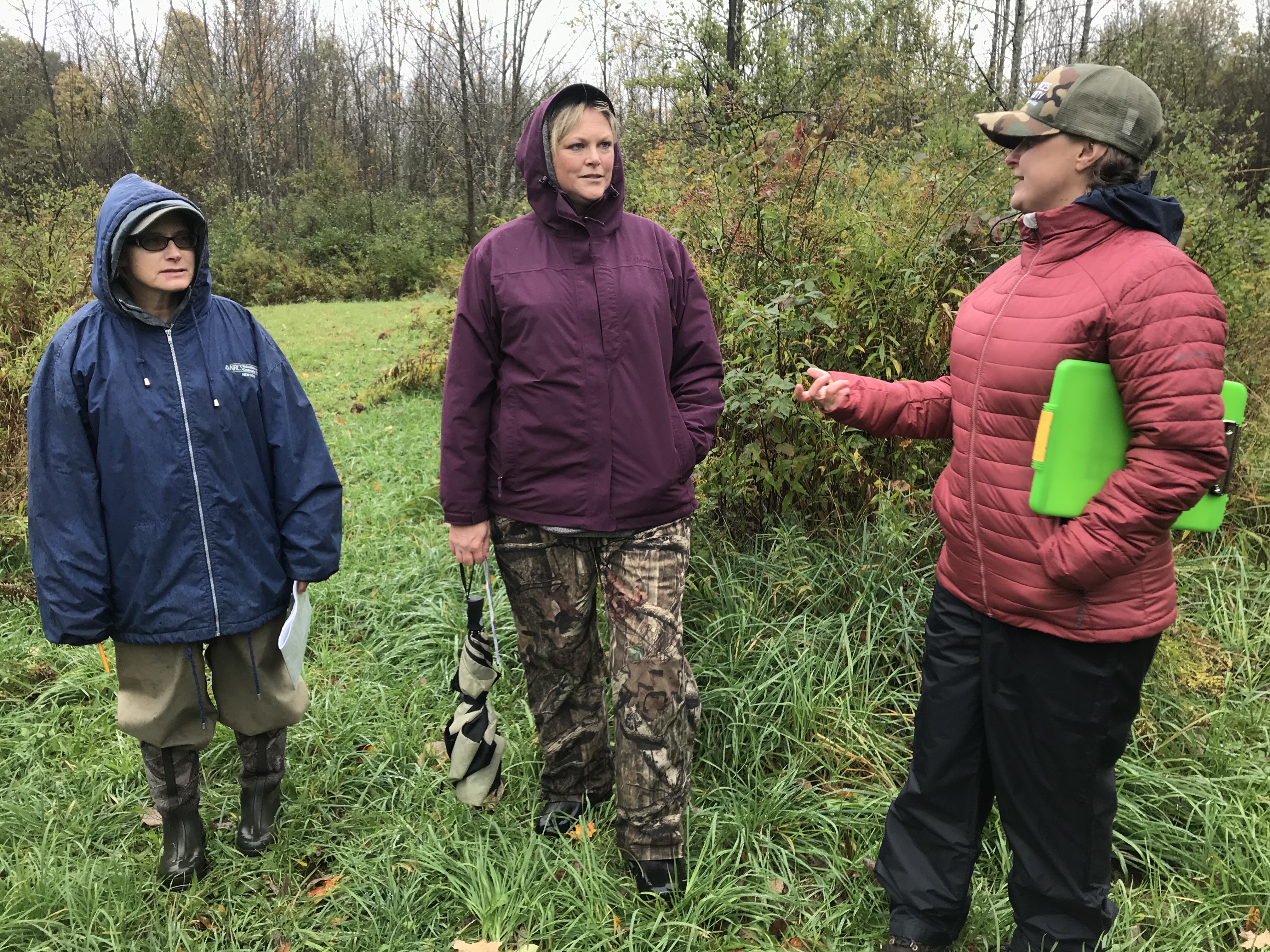 Three people stand outdoors in the rain. They are wearing heavy coats, and are in conversation.