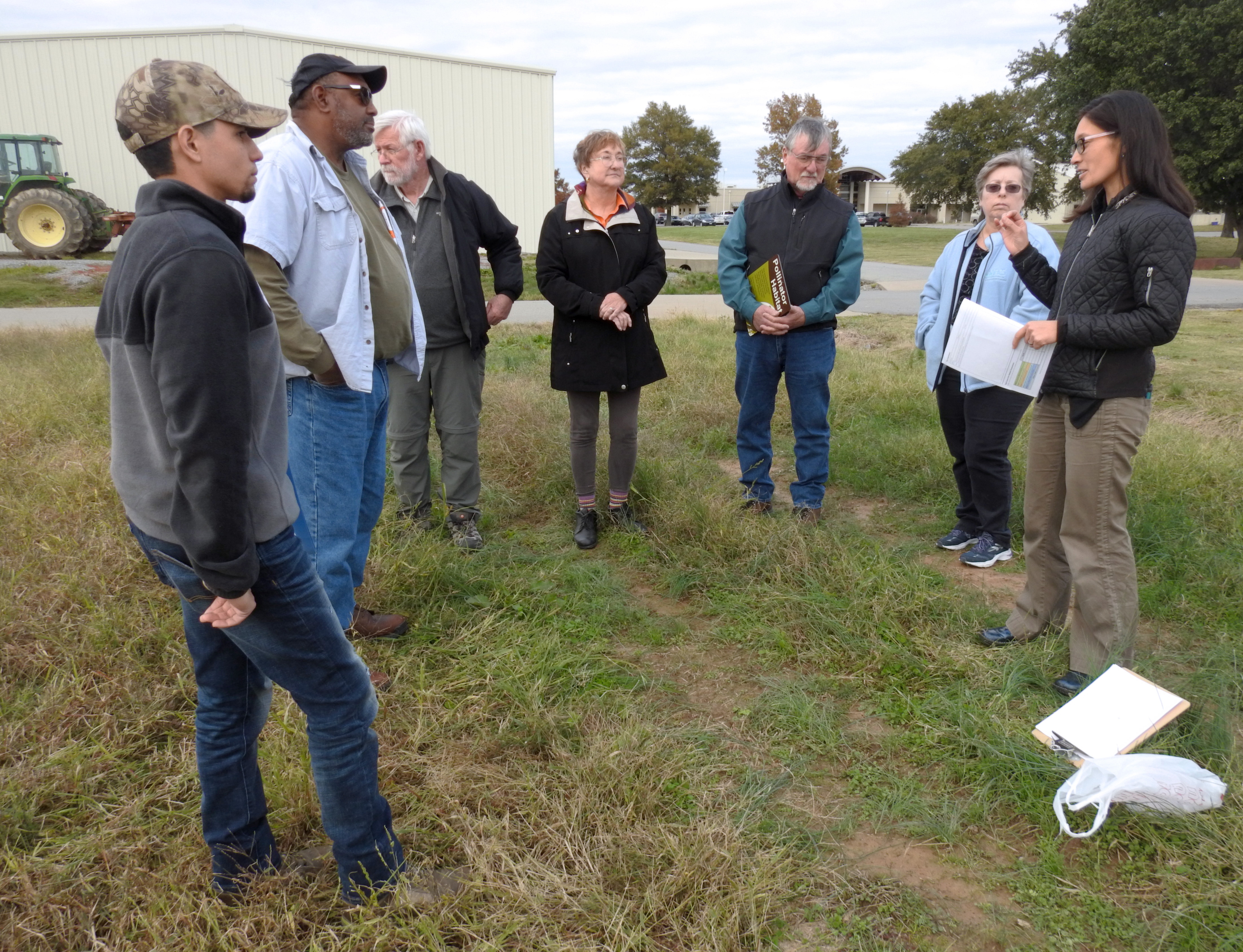 A group of people gathers around a woman (Thelma), who is speaking in an outdoor setting near a barn and tractor.