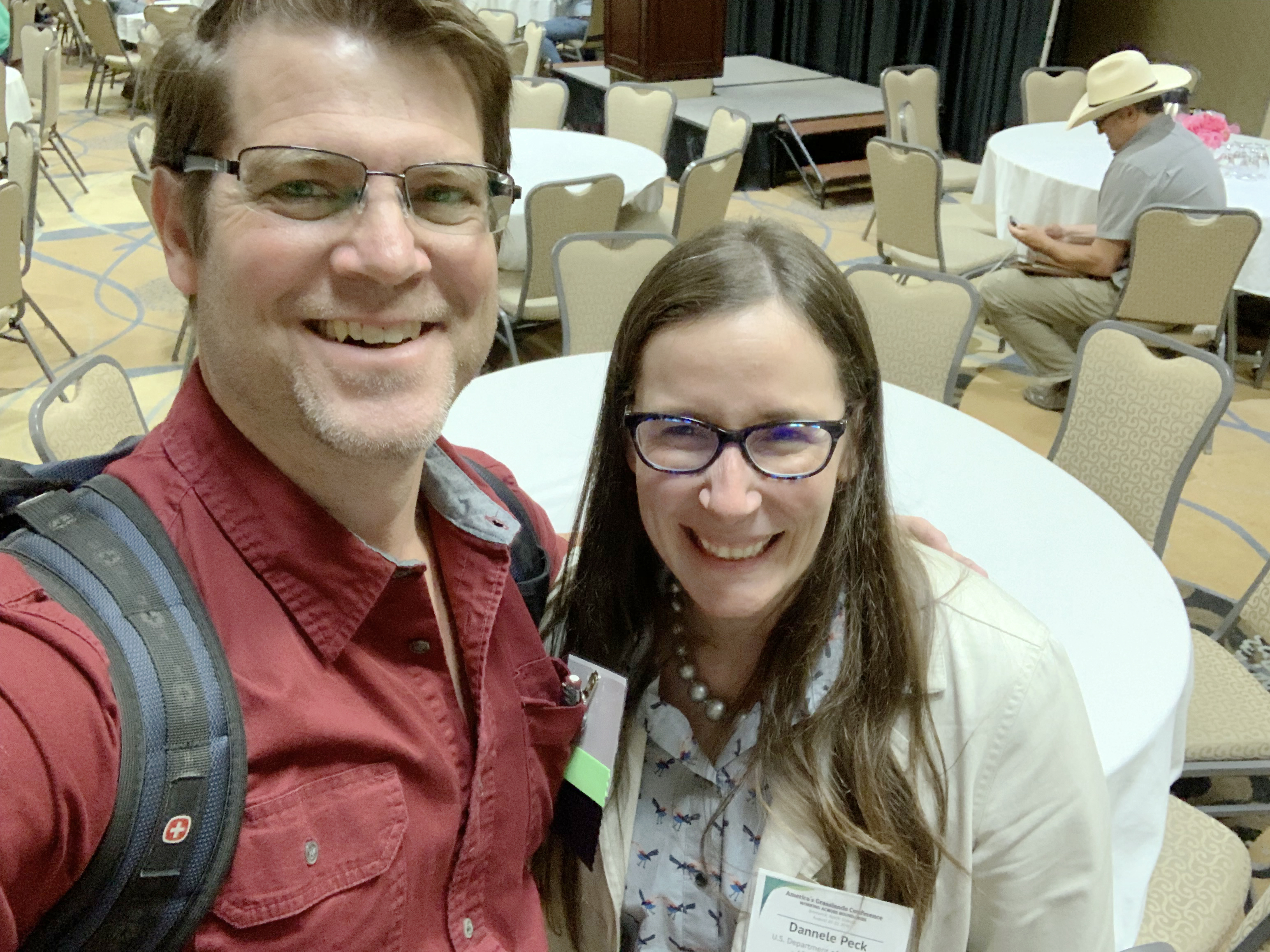 Two smiling people pose for a selfie in a room filled with tables.