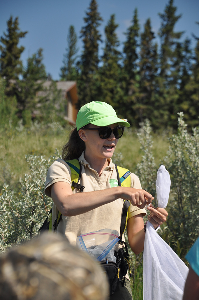 In an open area on a sunny day, a person in a baseball cap talks to a crowd while holding a white net.