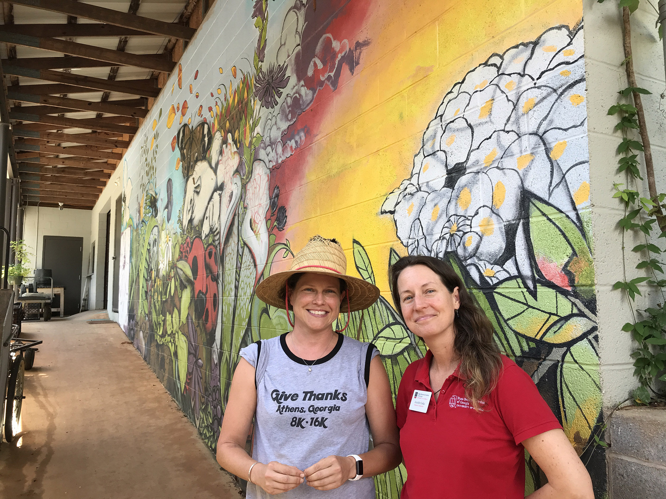 Two women in grey and red shirts stand in front of a colorful mural that shows flowers and insects at the State Botanical Garden of Georgia