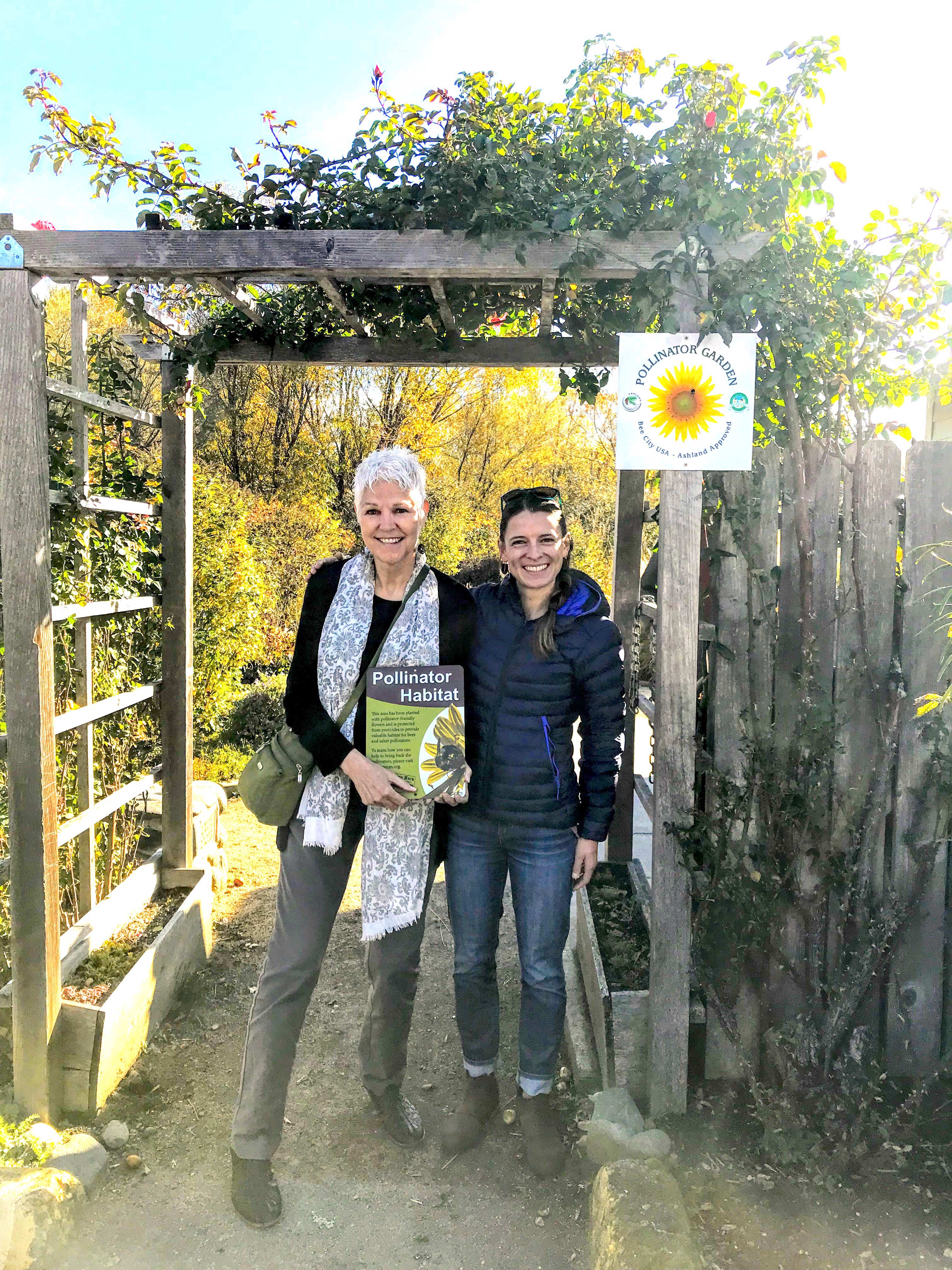 A tall woman with short, white hair who is holding a Xerces pollinator habitat sign stands next to a woman with dark hair and a dark down coat, in the entrance to a garden.