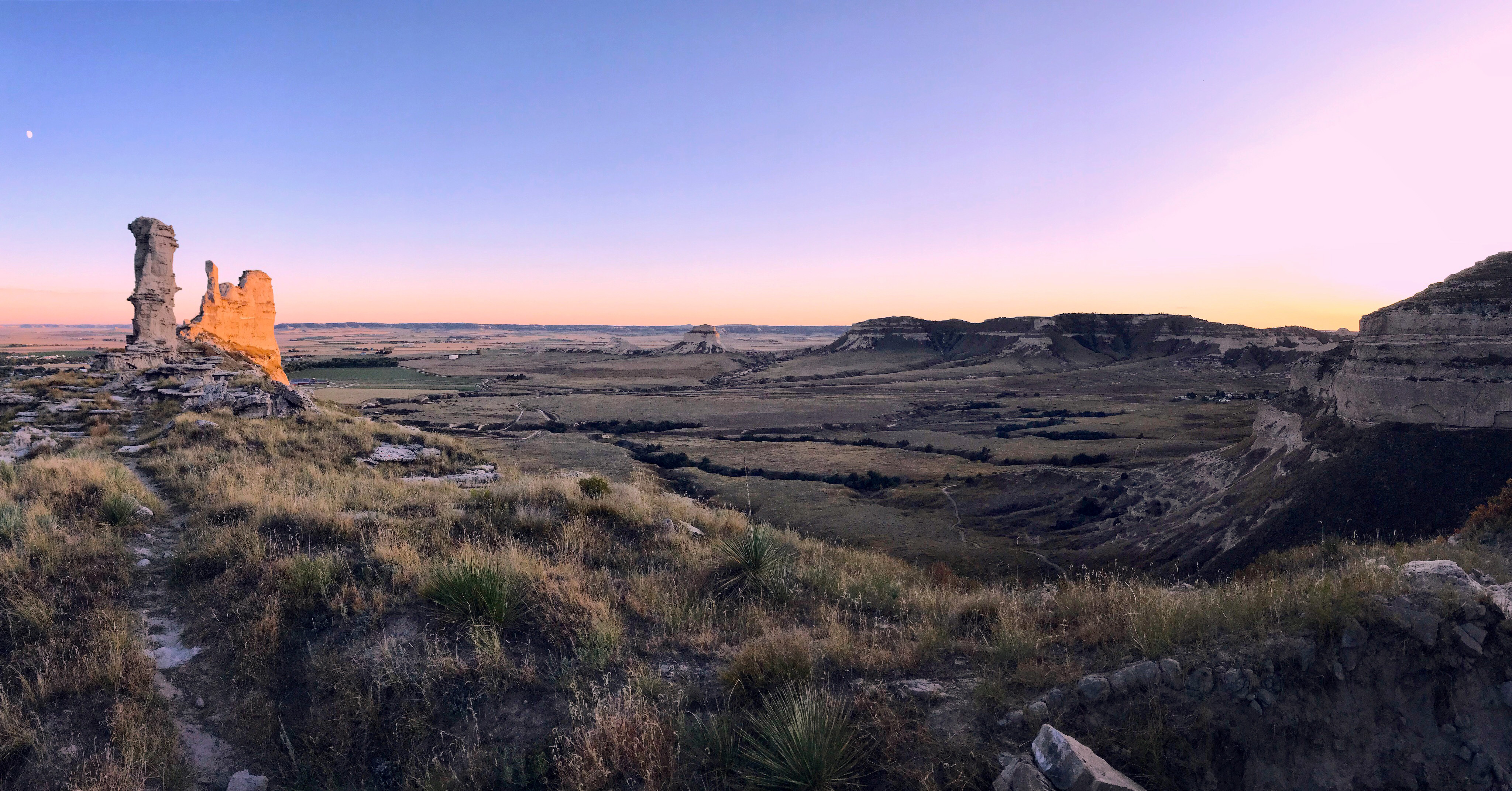 A dramatic landscape at dusk, with a thin rim of orange along the horizon. On the left are two tall rock formations, one shaped like a tower, and one similar to a trapezoid.