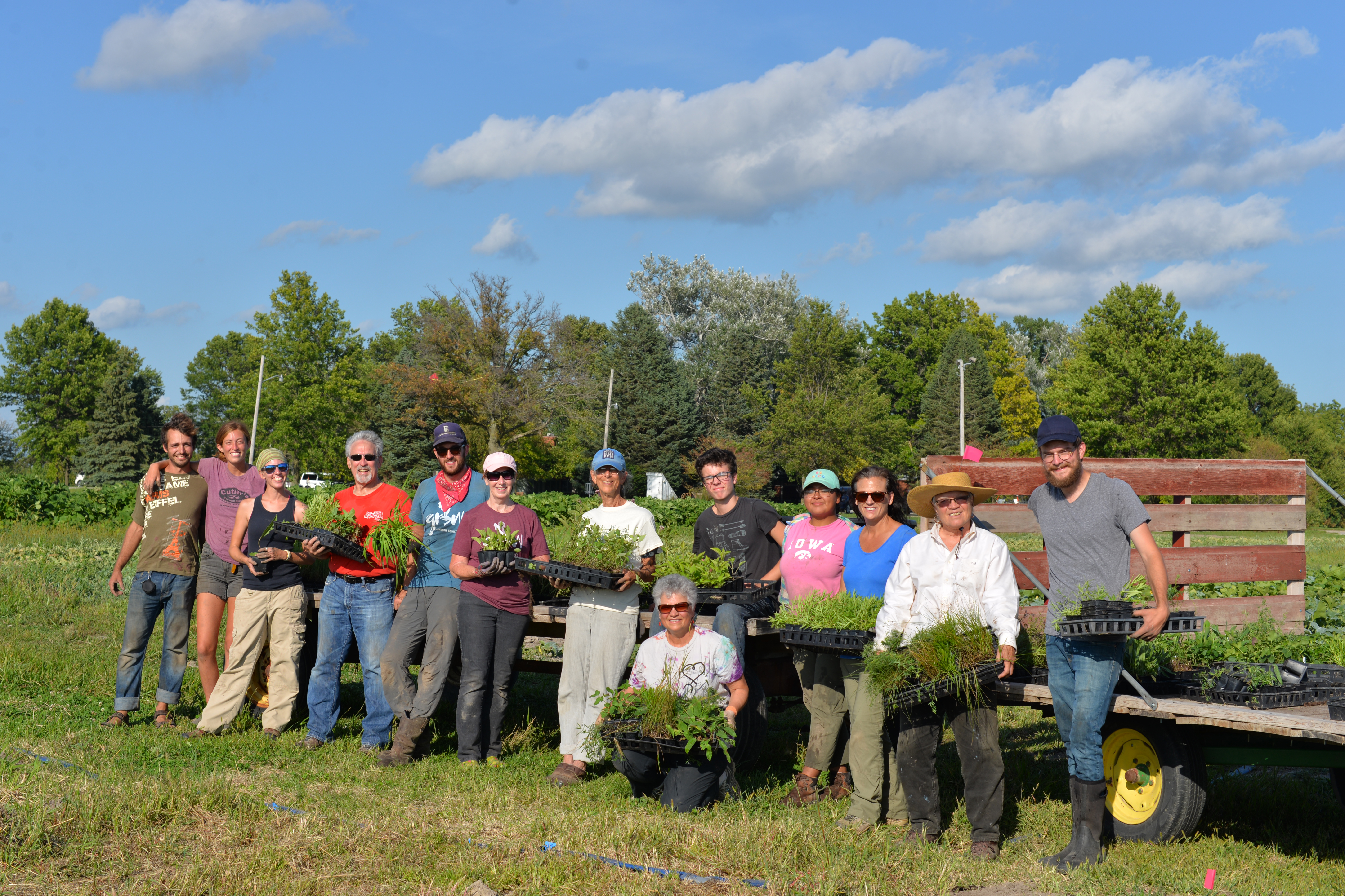 A group of smiling people of a diversity of ages, races, and genders stands next to a flatbed truck. Nearly everyone is holding a plant.