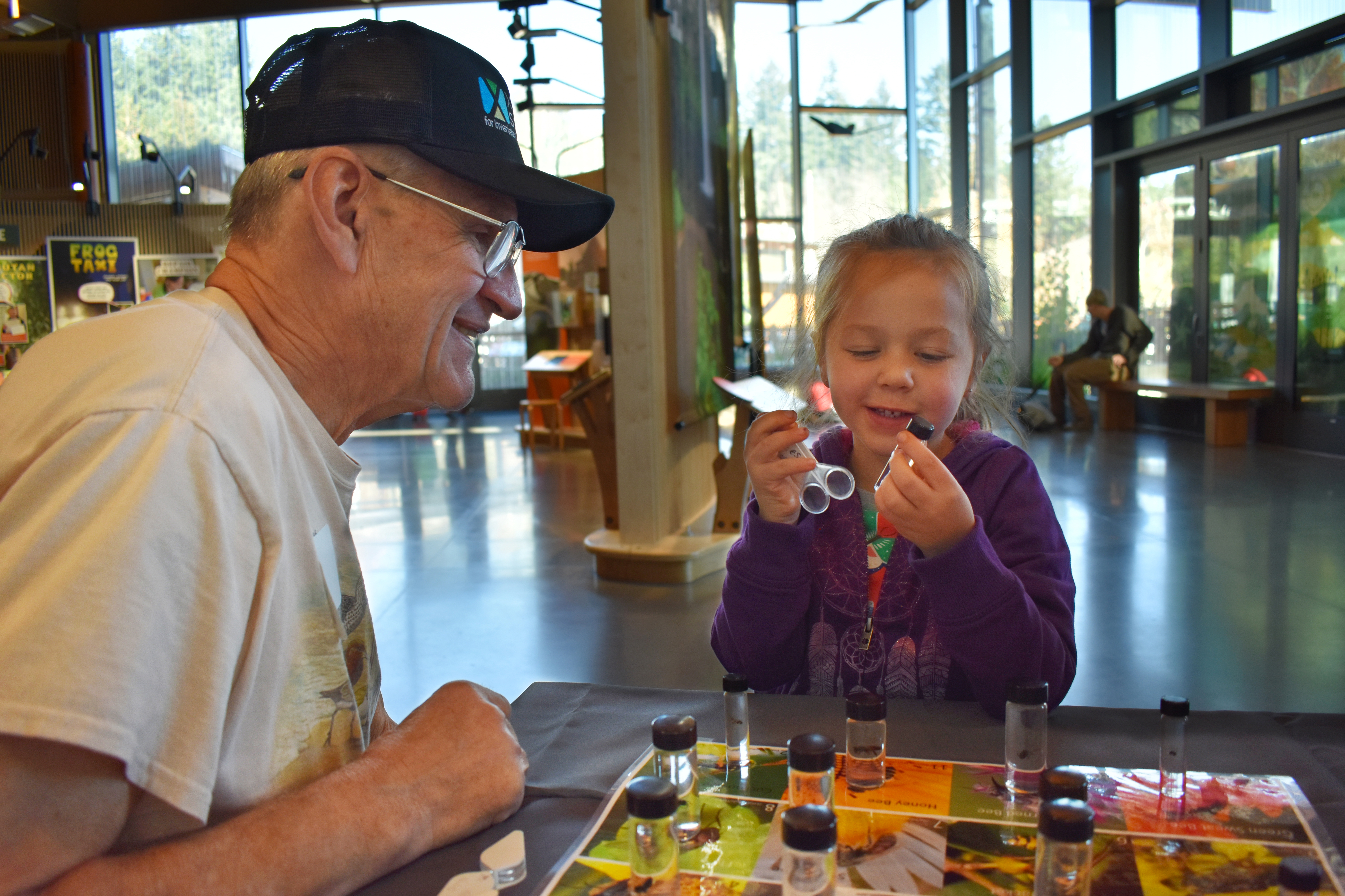 A Xerces volunteer talking with a little girl about bees at a table as she hols a magnifying glass.