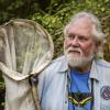 Robert Michael Pyle, who founded the Xerces Society in 1971. He has grey hair and beard, and is wearing a blue denim shirt over a blue t-shirt with a black-and-yellow swallowtail butterfly design -- and is holding his butterfly net.