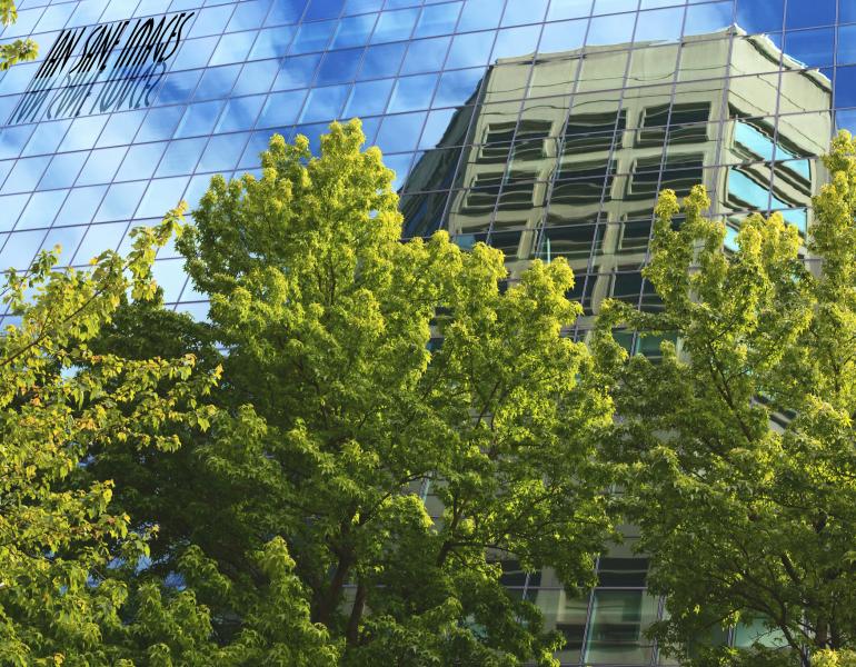 Bright green trees are shown alongside a wall of windows, reflecting a blue sky and a tall building.