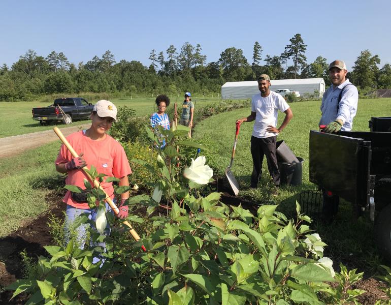 A group of smiling people poses with their shovels and other implements by a flowering hedgerow.