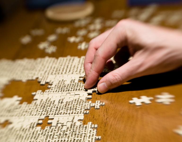 Pieces of a puzzle are being assembled on a dark, wooden table. A hand moves the pieces.