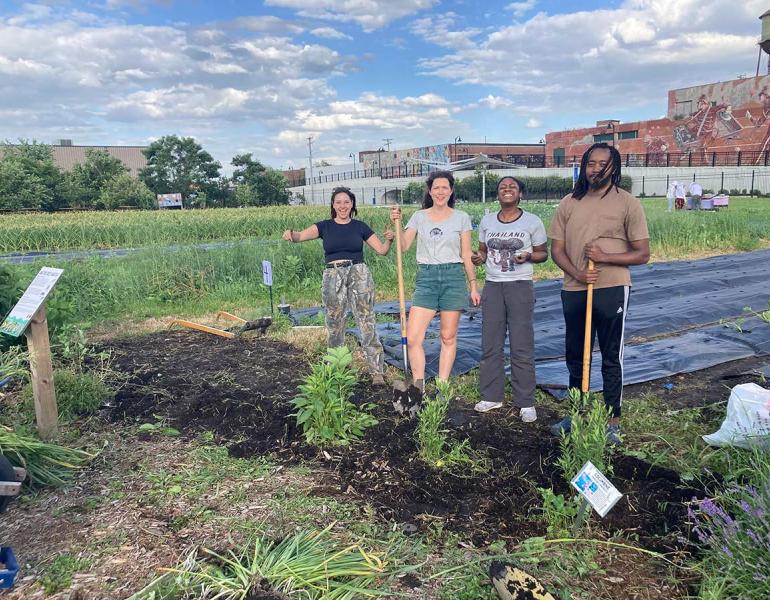  Four people, two white and two black, holding shovels and smiling while standing in front of several freshly planted native flowers. Behind them tarps protect a small plot of crops. 