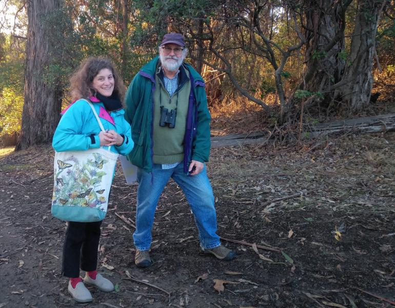 A man and a woman stand nest to each other in front of a grove of old trees.