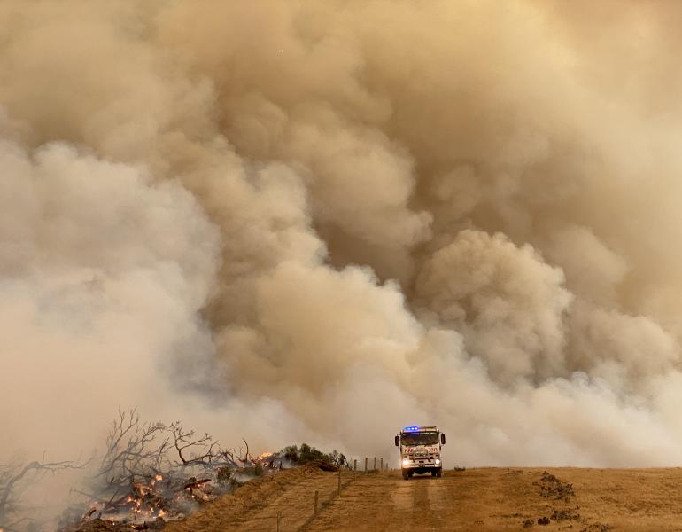 Fire truck drives across a burning landscape, with the sky full of brown smoke