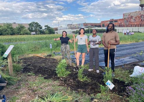  Four people, two white and two black, holding shovels and smiling while standing in front of several freshly planted native flowers. Behind them tarps protect a small plot of crops. 