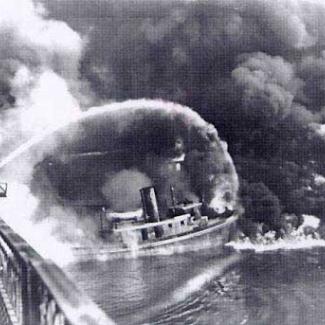 A firefighter sprays water from a bridge onto the burning Cuyahoga River in this historical photo, in black and white..