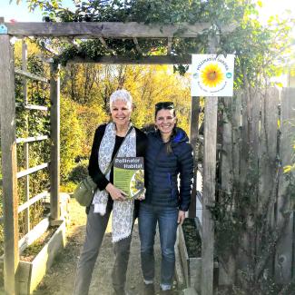 A tall woman with short, white hair who is holding a Xerces pollinator habitat sign stands next to a woman with dark hair and a dark down coat, in the entrance to a garden.