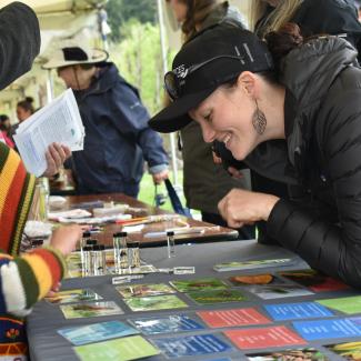 A smiling woman wearing a black coat and a black Xerces Society hat leans over a table. On the other side of the table, a kid with a colorful hoodie reaches for an assortment of small glass vials containing bee specimens. The woman and the kid are looking at the same vials and appear to be talking.