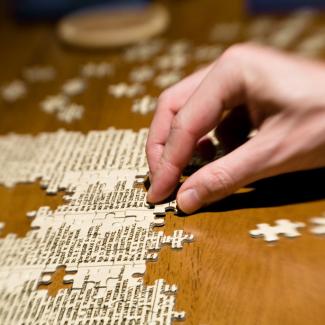 Pieces of a puzzle are being assembled on a dark, wooden table. A hand moves the pieces.