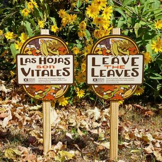 Xerces yard signs in front of a home garden, with fallen leaves on the ground. One reads "Leave the Leaves", while the second, in Spanish, reads "Las Hojas son Vitales".