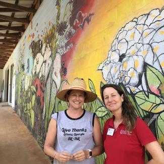 Two women in grey and red shirts stand in front of a colorful mural that shows flowers and insects at the State Botanical Garden of Georgia