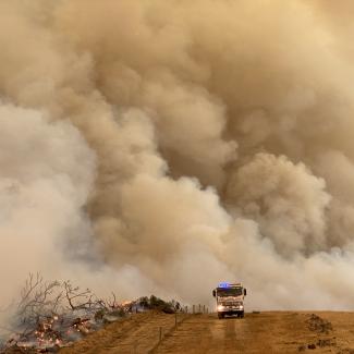 Fire truck drives across a burning landscape, with the sky full of brown smoke