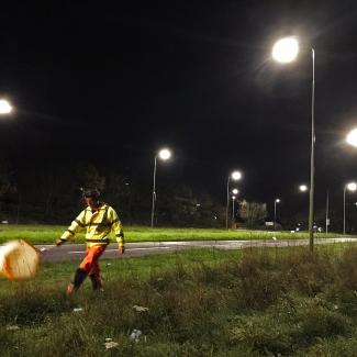 A person wearing fluorescent clothing and a headlamp swings a net alongside a roadway lit by streetlamps at night.