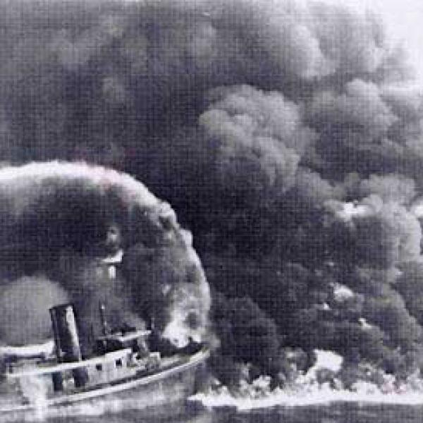 A firefighter sprays water from a bridge onto the burning Cuyahoga River in this historical photo, in black and white..